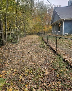 A gravel driveway lined with metal fencing leading to a blue-gray house surrounded by autumn trees with fallen leaves